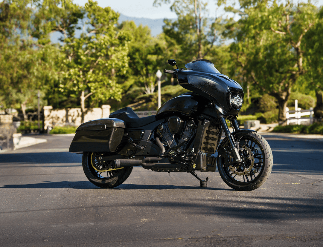 A sleek, all-black Indian brand cruiser motorcycle parked on an asphalt driveway with lush green trees in the background